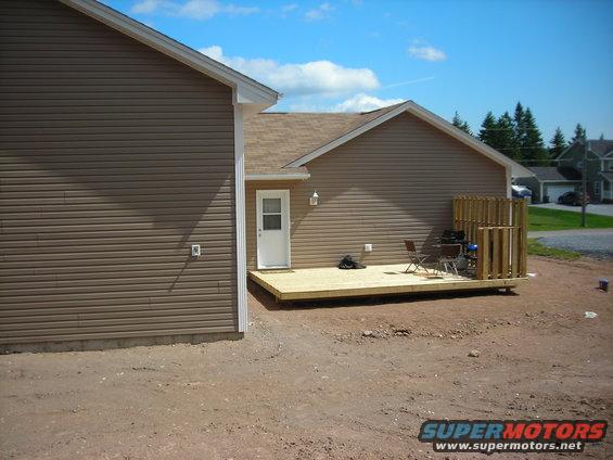 Exterior view of a house with brown siding, a wooden deck, and a grill. A blue sky with clouds is visible