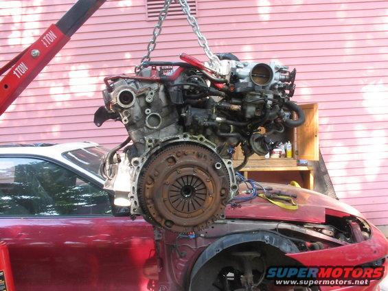 Red car with engine removed, engine suspended by chains from a red crane