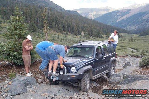 People working on a dark Jeep Liberty stuck on a rocky mountain trail