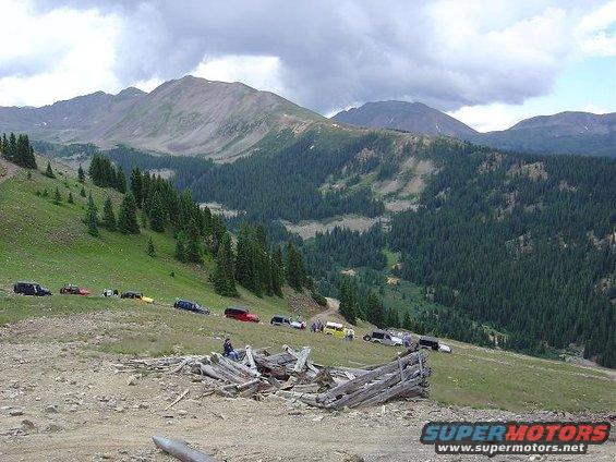 Multiple SUVs driving on a mountain road with pine forests and peaks in the background