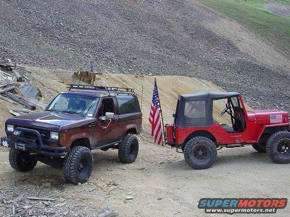 1986 Ford Bronco II and a red Jeep parked side-by-side on a dirt road with an American flag on the Jeep