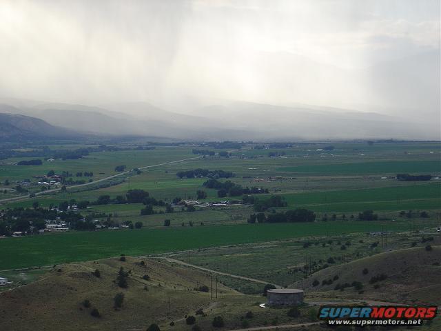 Overhead view of a vast rural landscape with rolling hills, green fields, and distant mountains under a cloudy, rainy sky