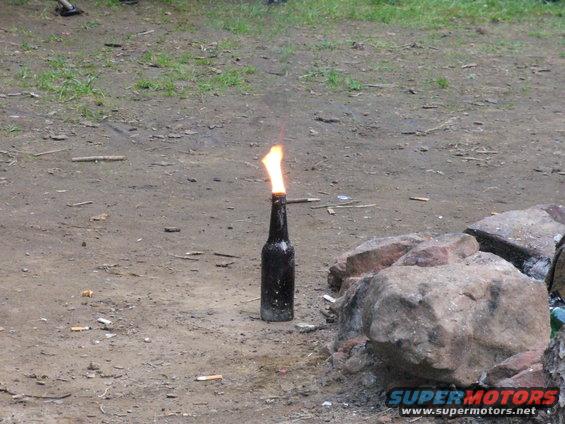 Bottle with flame on top, sitting on dirt ground next to rocks. Cigarette butts are scattered around