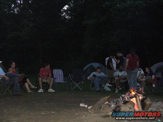 Group of people sitting in camping chairs around a campfire at night, with tents in the background