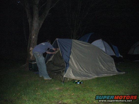 Person setting up a tent at night, with other tents in the background