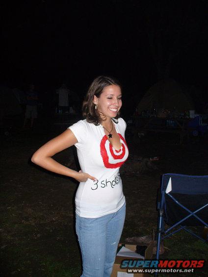 Woman in jeans and a white t-shirt with a red target graphic and '3 shots' text. Camping tents and chairs are visible in the background