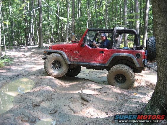 Red 2003 Jeep Wrangler with large tires driving through a muddy trail in a forest