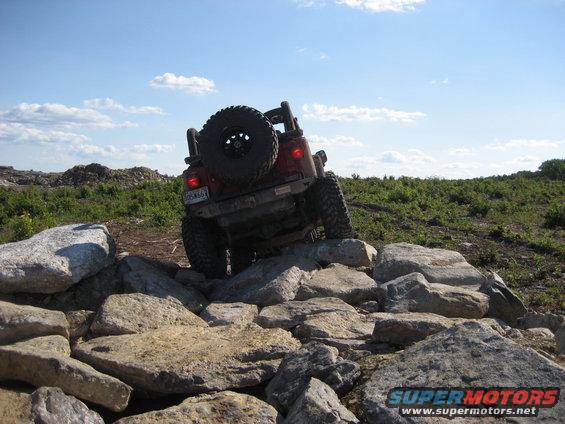 Red Jeep Wrangler climbing over large rocks on a dirt trail under a blue sky with clouds