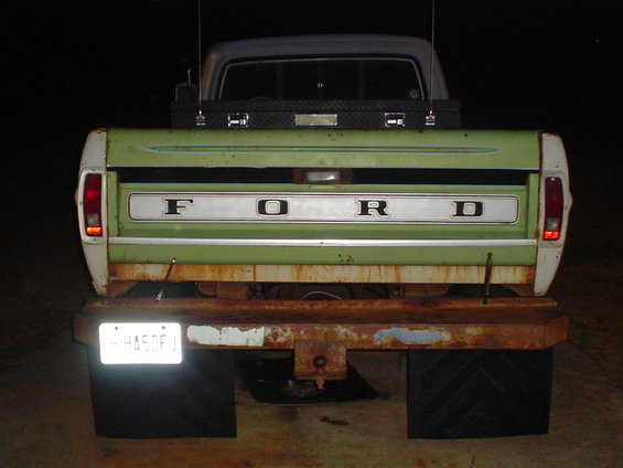Green and white Ford pickup truck tailgate with chrome trim and "FORD" lettering. Rusty bumper and black mud flaps visible