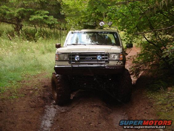 Cream-colored Ford Bronco with large mud tires driving on a muddy trail