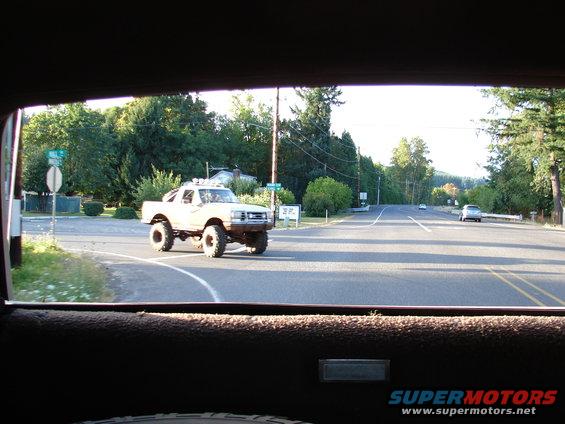 Lifted pickup truck with large tires turning at an intersection. Trees and power lines line the road