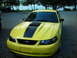 2003 Ford Mustang - A yellow 2003 Ford Mustang with black racing stripes is parked on a gravel surface near a body of water.