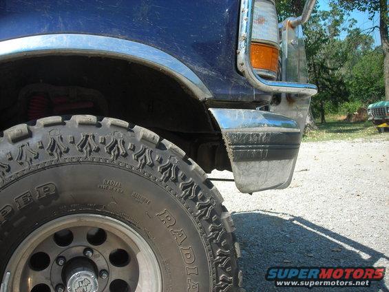 Blue Ford truck front fender and bumper with chrome trim, off-road tire, and a green truck in the background