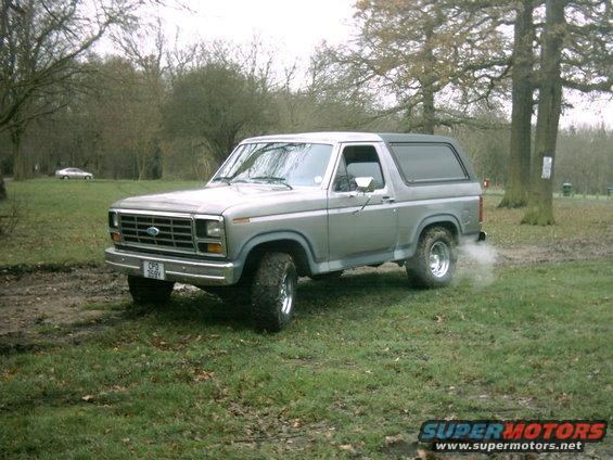 Silver Ford Bronco with aftermarket wheels and tires, seen from a 3/4 front angle in a grassy, muddy field