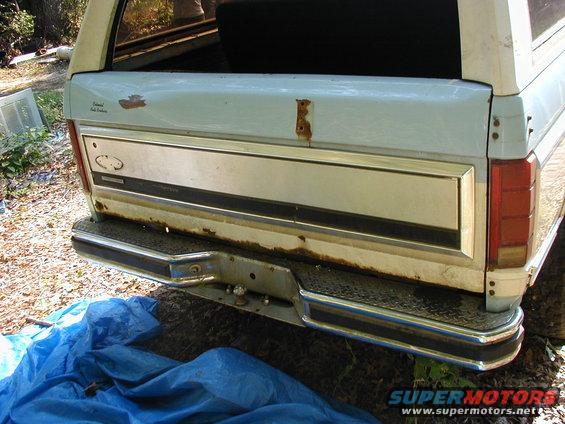 Rear of a light blue SUV with chrome bumper and tailgate trim, showing rust spots and a blue tarp