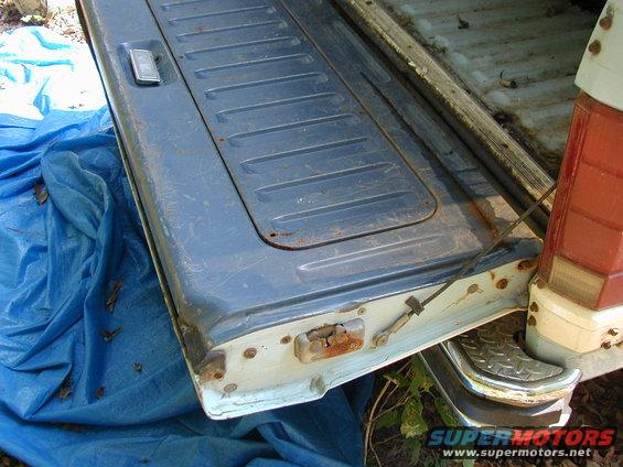 Blue tailgate with horizontal ridges and a chrome handle, resting on a blue tarp. Rusty latch mechanism visible