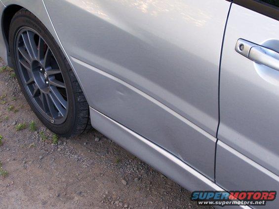 Silver car's side door and multi-spoke wheel. Ground is gravel and grass