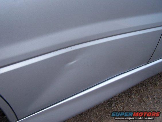 Silver car door with a noticeable dent near the body line. A thin chrome trim piece runs along the bottom edge