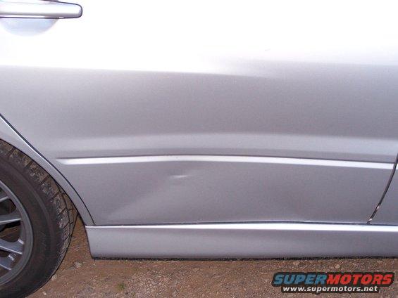 Silver car door with a dent near the bottom, showing a portion of the rear wheel and a door handle