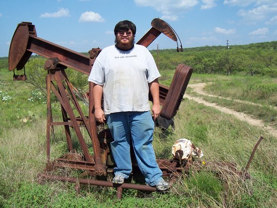 Man in a t-shirt that says "it's all muscle" stands in front of a rusty oil pumpjack in a grassy field