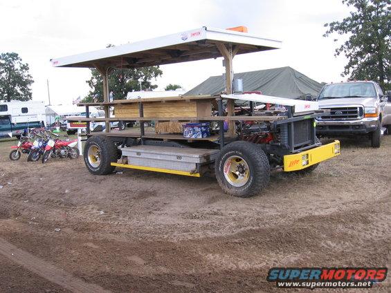 Custom vehicle with a wooden bar structure and canopy, parked next to a pickup truck and motorcycles