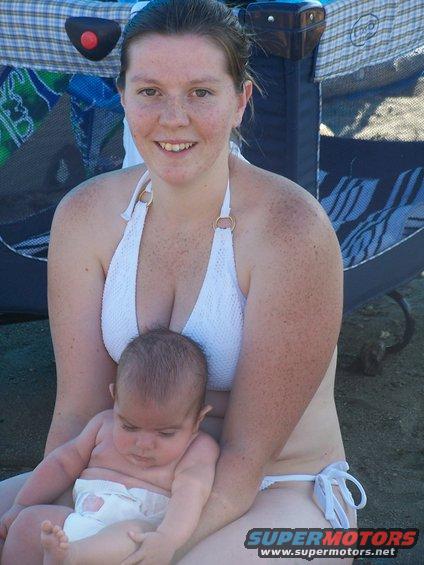 Woman in white bikini holding a baby on the beach, with a playpen behind them. Supermotors.net logo visible