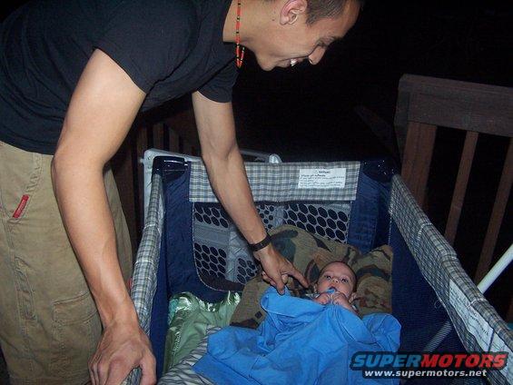 Man leans over a playpen at a baby covered with a blue blanket. Text on playpen and SuperMotors logo visible
