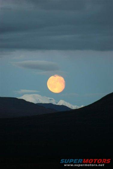 Full moon rises over snow-capped mountains and dark hills under a cloudy sky