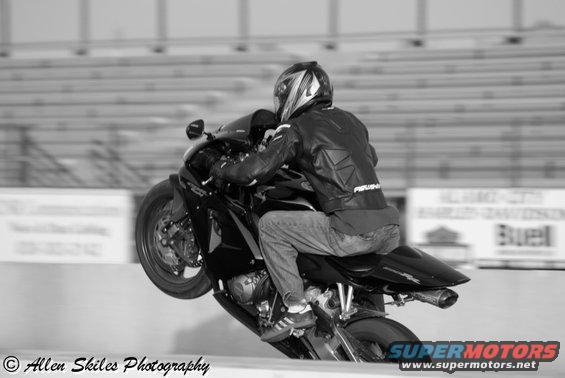 Motorcyclist on a black sportbike doing a wheelie on a track. Rider wears a helmet and leather jacket