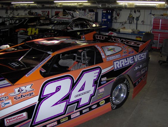 Race car number 24 with orange and black paint scheme, parked in a garage with other cars