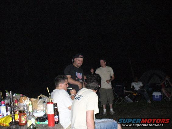 Group of people at a campsite at night, with drinks and a fire extinguisher on a table
