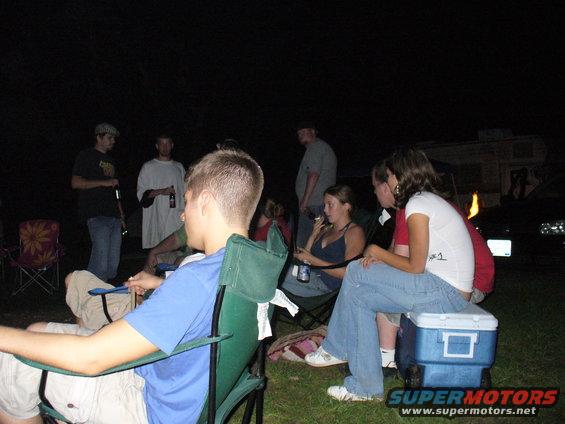 People gathered outdoors at night, some sitting in chairs, with a trailer and a dark vehicle in the background
