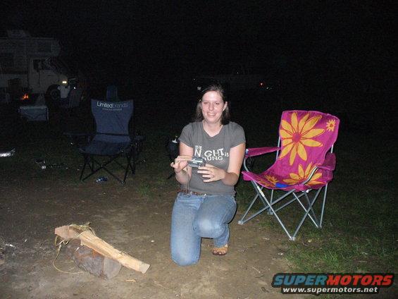 Woman kneeling in front of a pink floral chair, holding a camera, with a camper van in the background at night