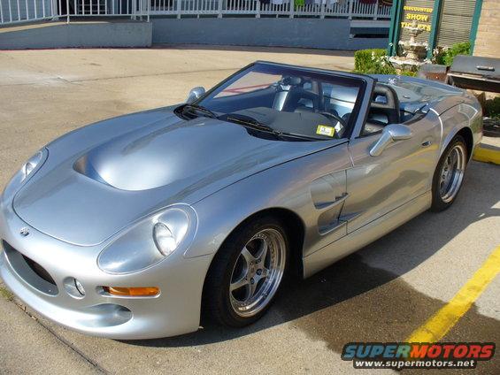 Silver convertible sports car with a hood scoop and side vents, parked on asphalt with yellow lines