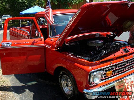 Red 1965 Ford F-100 pickup truck, Custom Cab trim, hood open showing engine. American flag on fender
