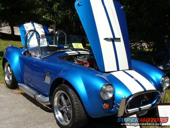 Blue AC Cobra replica, hoods open, showing engine and interior. White racing stripes on hood and rear deck