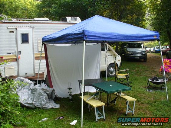 Camper trailer, white van under blue canopy, picnic table, chairs, and other vehicles in background