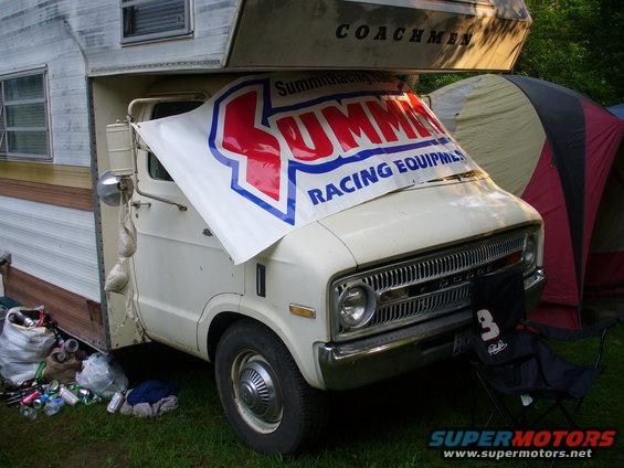 White RV with a large banner covering the windshield, parked on grass with trash and a tent nearby