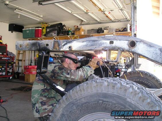 Guy in camo welding a vehicle frame in a garage. Big tires visible