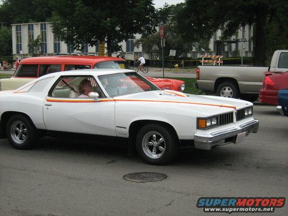 White 1975 Pontiac Grand Am coupe with orange and yellow side stripes, chrome wheels, and a woman driving
