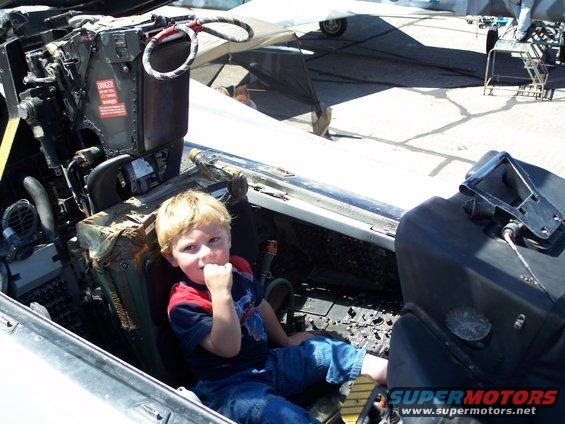 Kid sits in the cockpit of a jet fighter, looking at the camera