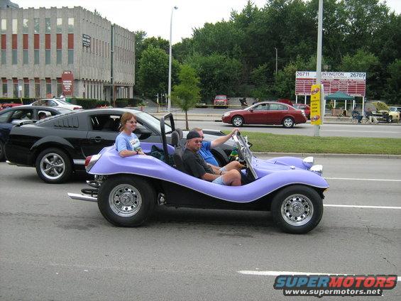 Purple dune buggy with two people driving down a street, black Mustang next to it