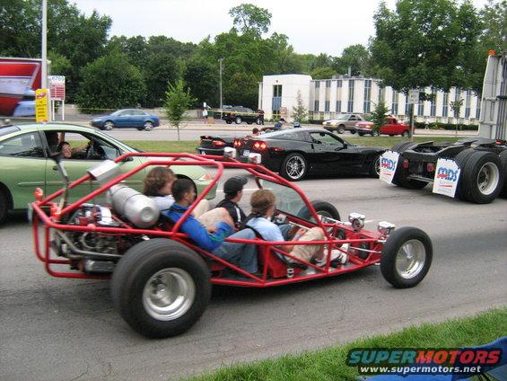 Red dune buggy with four people driving on a street, with other cars and buildings in the background