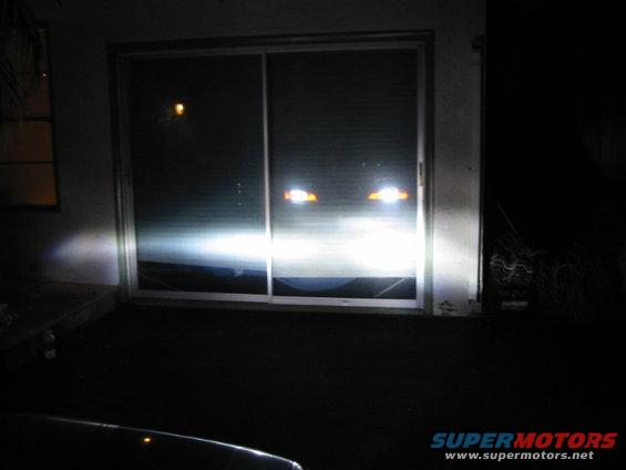 Car headlights shining brightly at a sliding glass door at night. Supermotors.net logo visible
