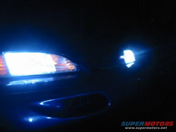Front of a blue car with bright blue headlights on at night. A side mirror is visible on the right