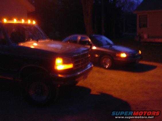 Dark sedan, likely a 1995 Ford Crown Victoria, parked at night with its headlights on