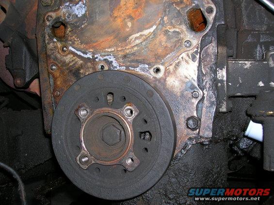 Engine timing cover with rust and oil, showing a crankshaft pulley with bolt pattern