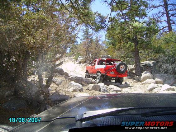Red SUV with spare tire on back driving on rocky trail. Another white SUV is further up the path