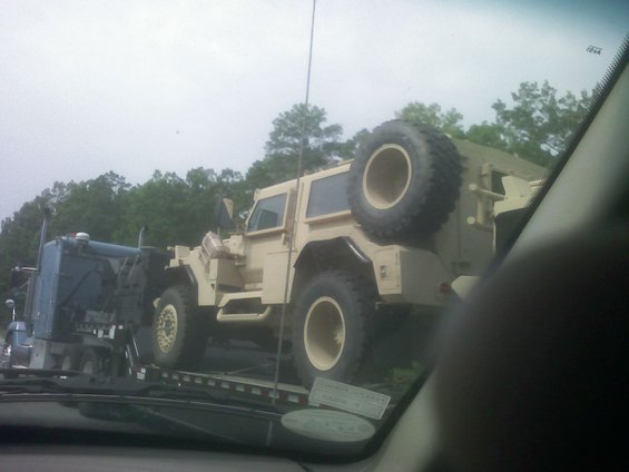 Tan military vehicle with a spare tire mounted on the rear, being transported on a flatbed truck