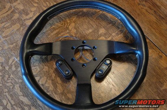 Black steering wheel with white stitching and a black hub, two cruise control button pods